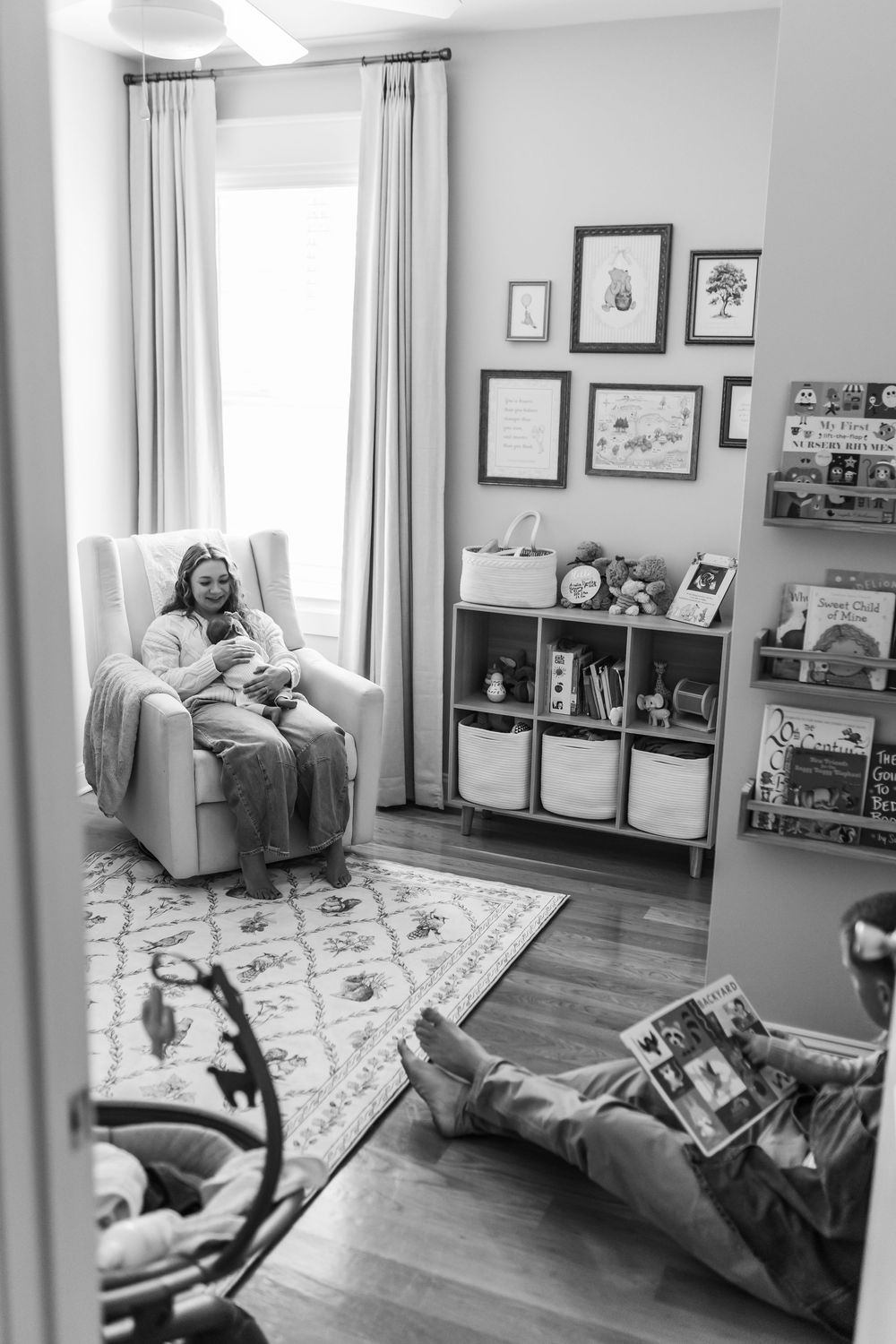 Black and white photo of mother in glider holding baby while partner reads on floor in cozy nursery.
