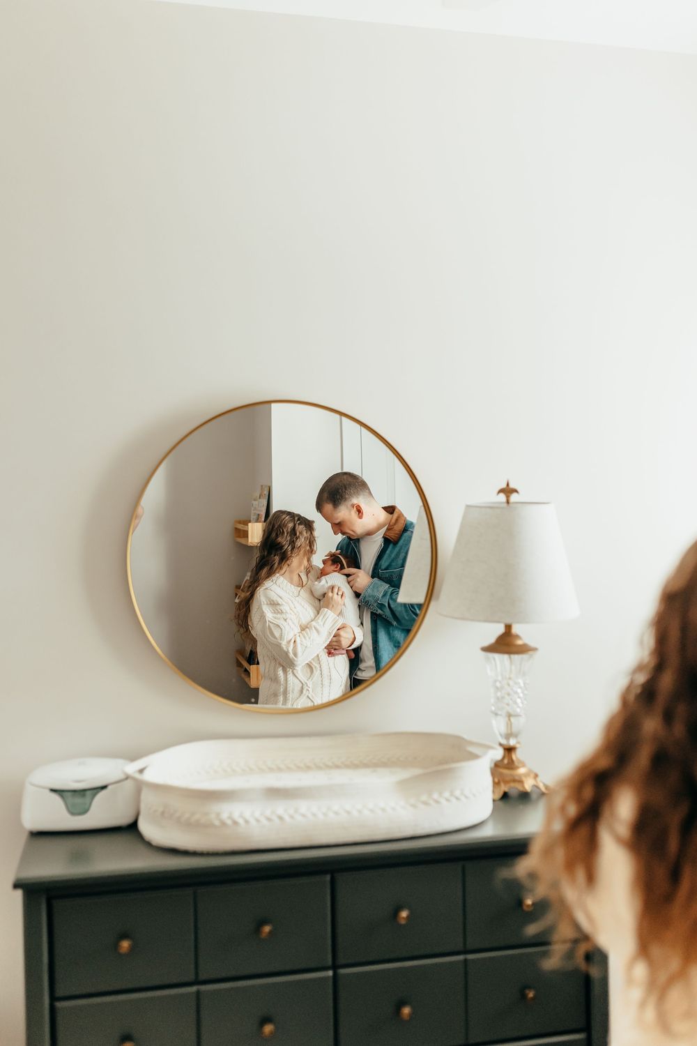 Couple holding newborn baby reflected in round gold mirror above green dresser with changing pad and lamp.