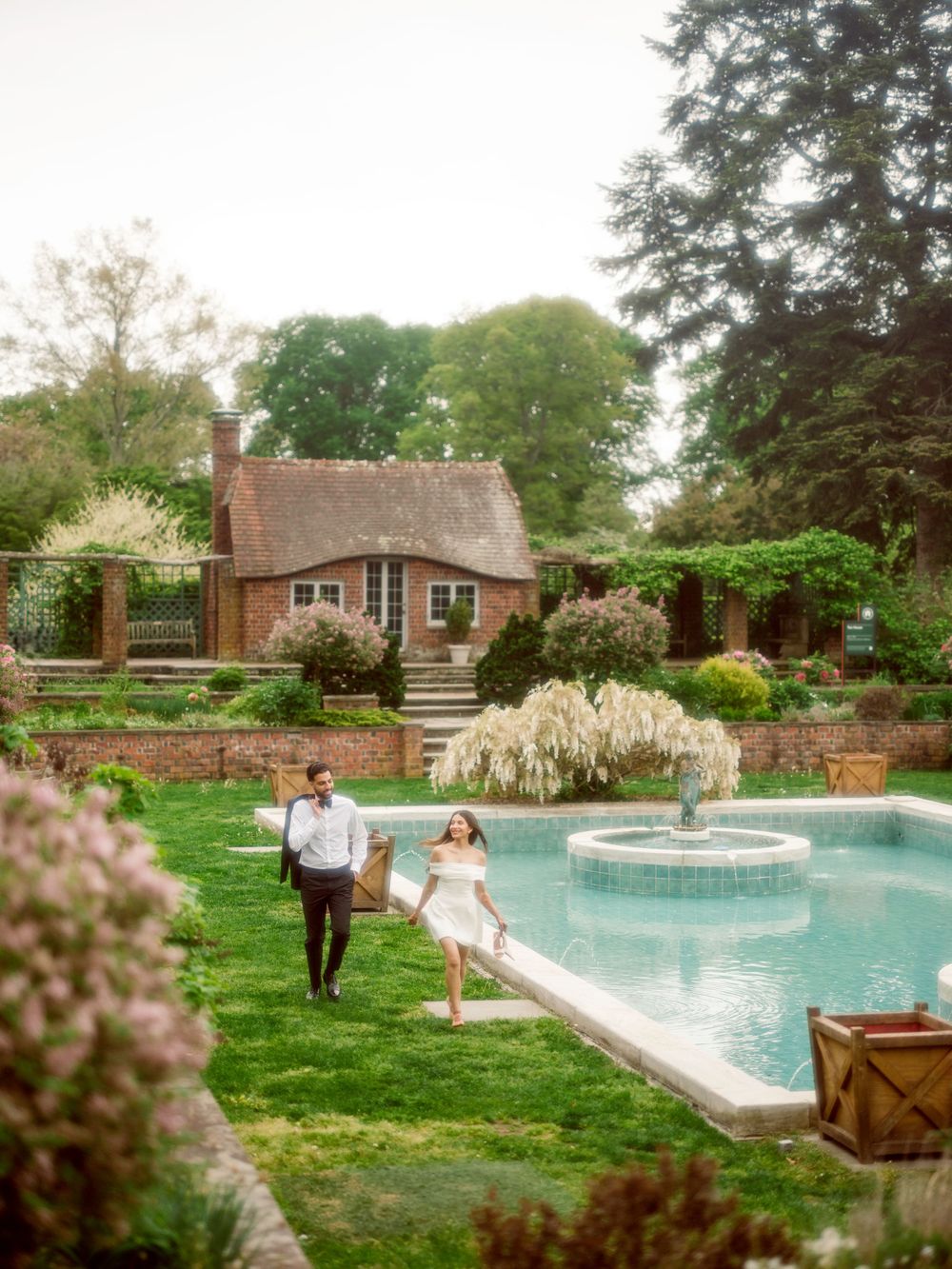 A couple walks along a lush garden path beside an elegant swimming pool with a fountain, next to a charming brick cottage.
