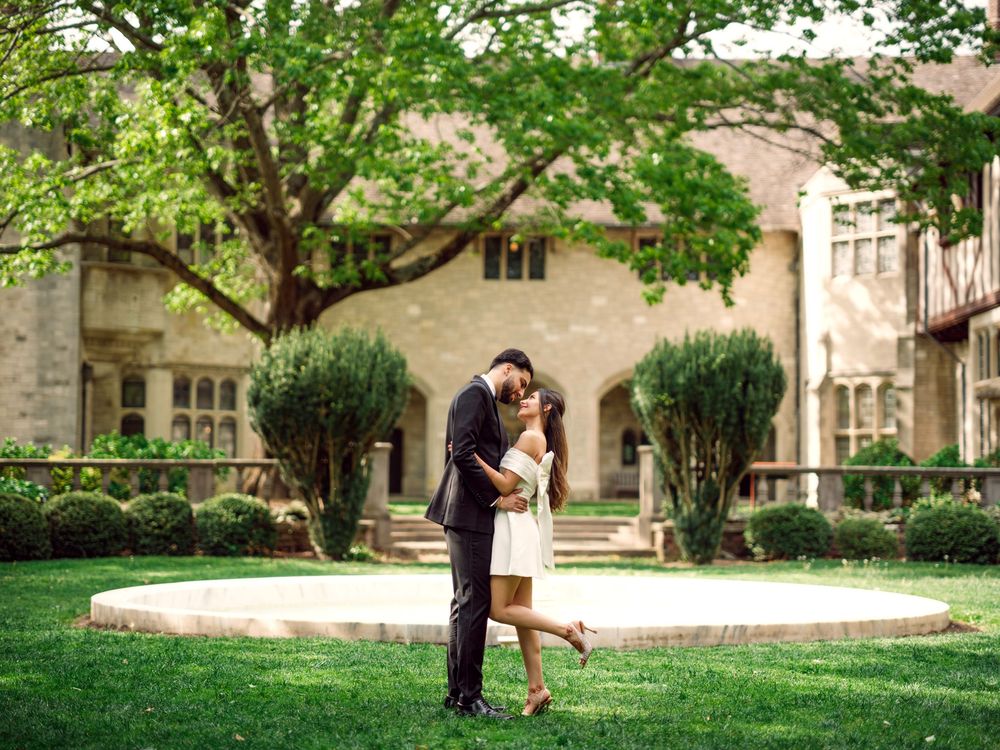 A couple embraces on a manicured lawn in front of a grand stone building with Gothic architecture and green trees.