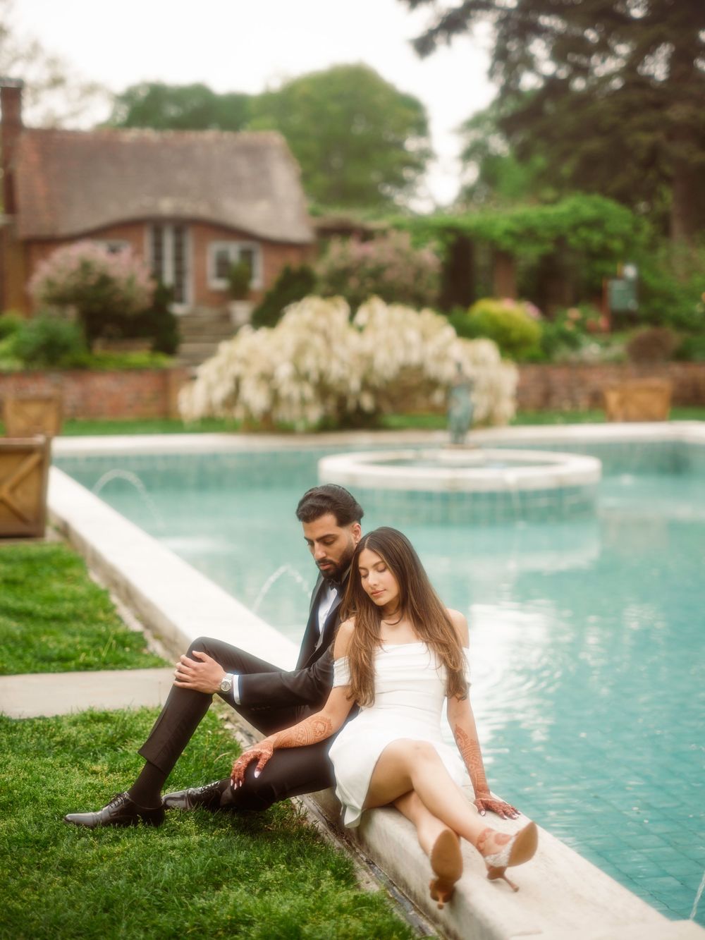 A couple sits together on the edge of a swimming pool in a garden setting with blooming white flowers and a house in background.