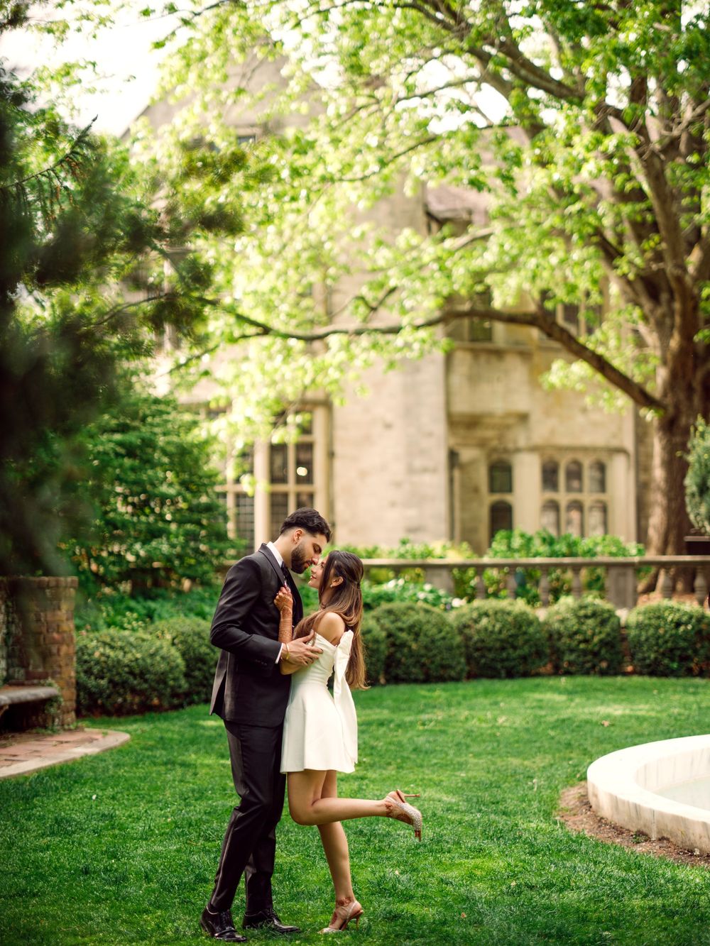 Romantic couple embraces on manicured lawn in front of elegant stone building with lush green trees.