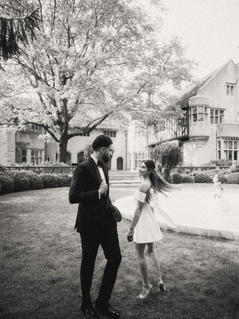 Two figures share a playful moment on a lawn with blooming trees and a stately building in the background.