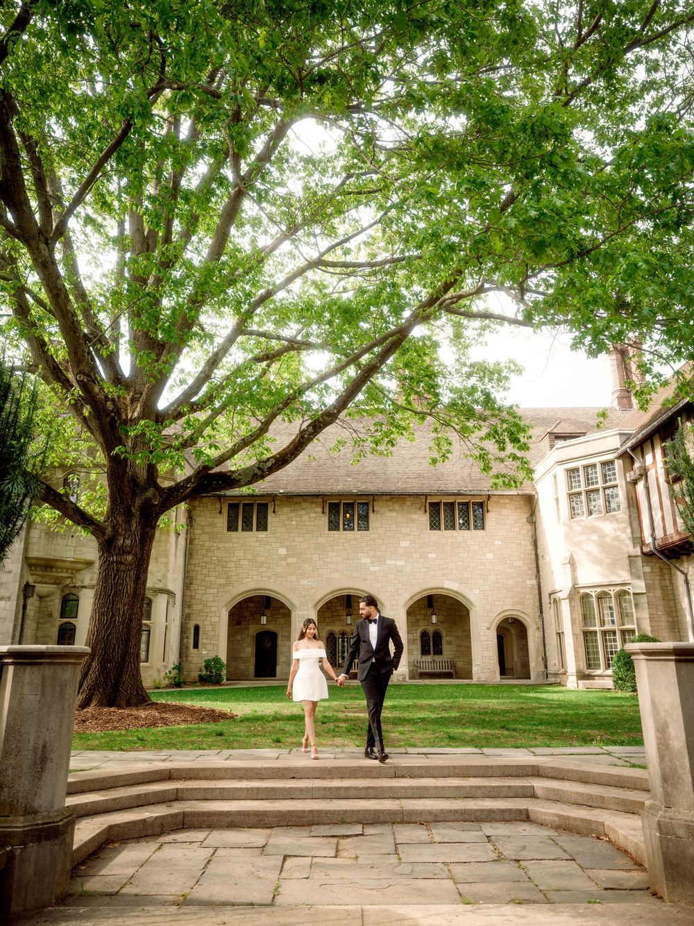 A couple stands on stone steps in front of a collegiate Gothic architecture building with arches and green trees overhead.