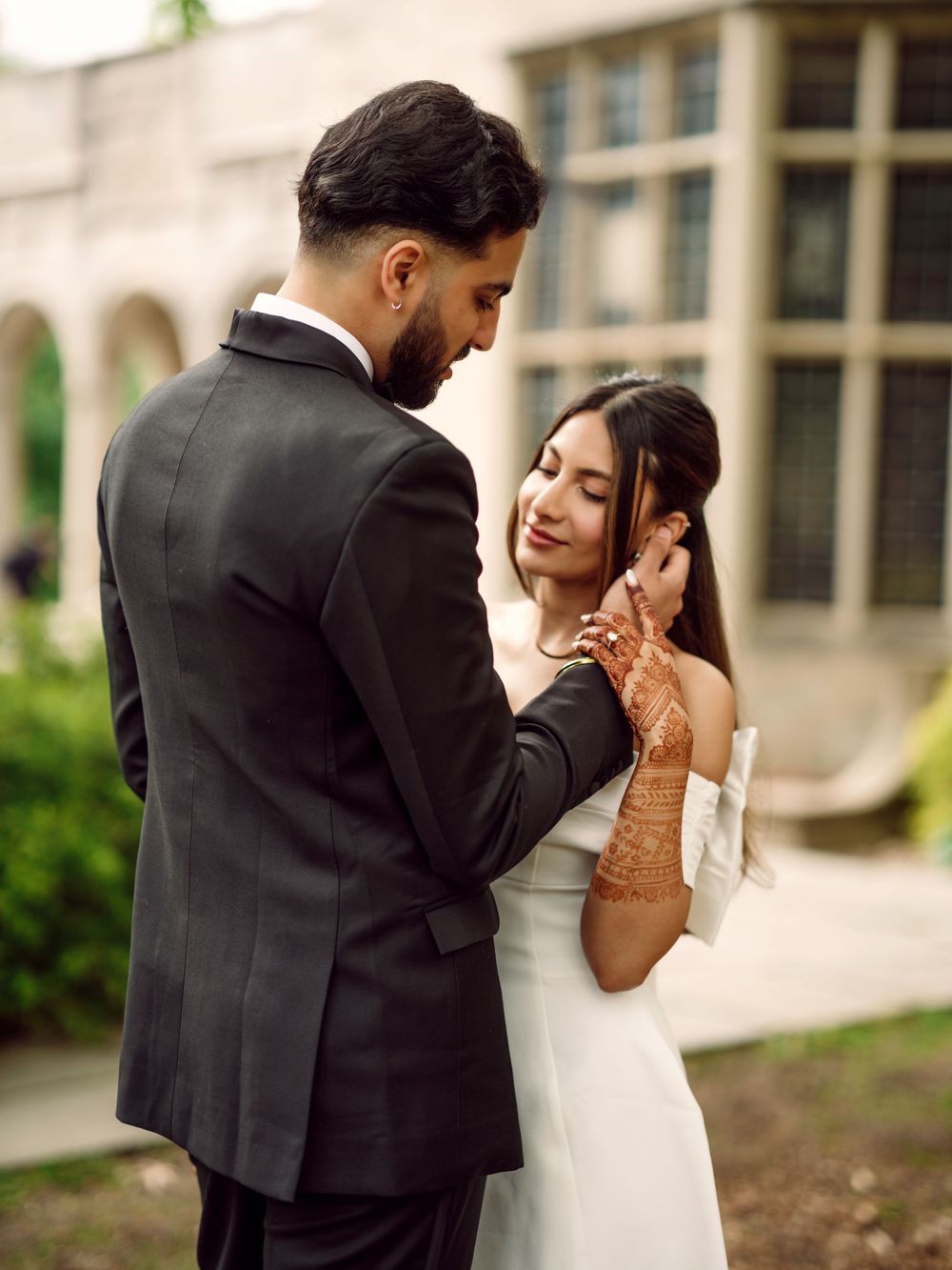 A bride in a white dress with henna decorated hands shares an intimate moment with her groom in a dark suit outdoors.