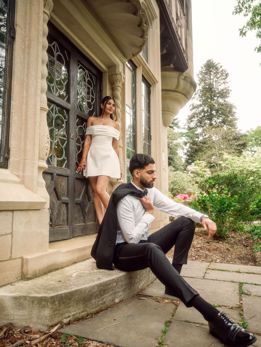 A couple poses elegantly on stone steps outside a historic building, with the woman in white and the man in formal attire.