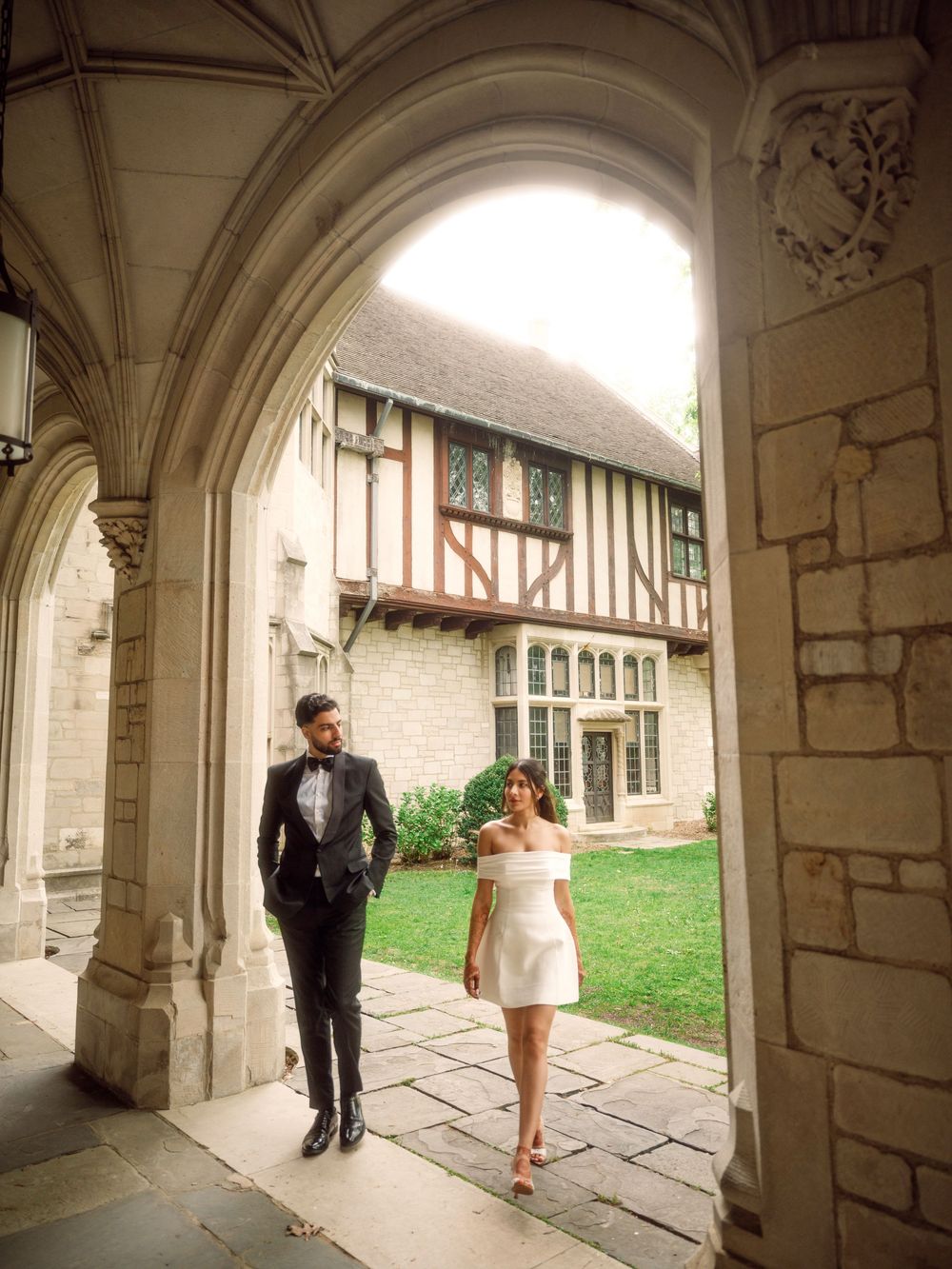 A couple in formal attire walks through a Gothic stone archway leading to a Tudor-style building.