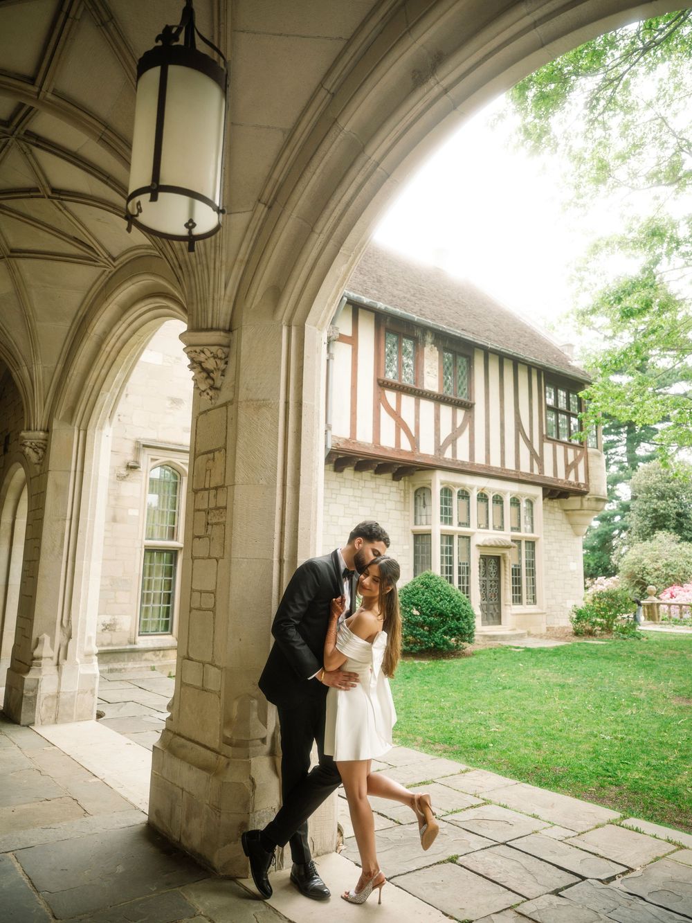 A romantic couple stands beneath a Gothic stone archway next to a Tudor-style building on a grassy lawn.
