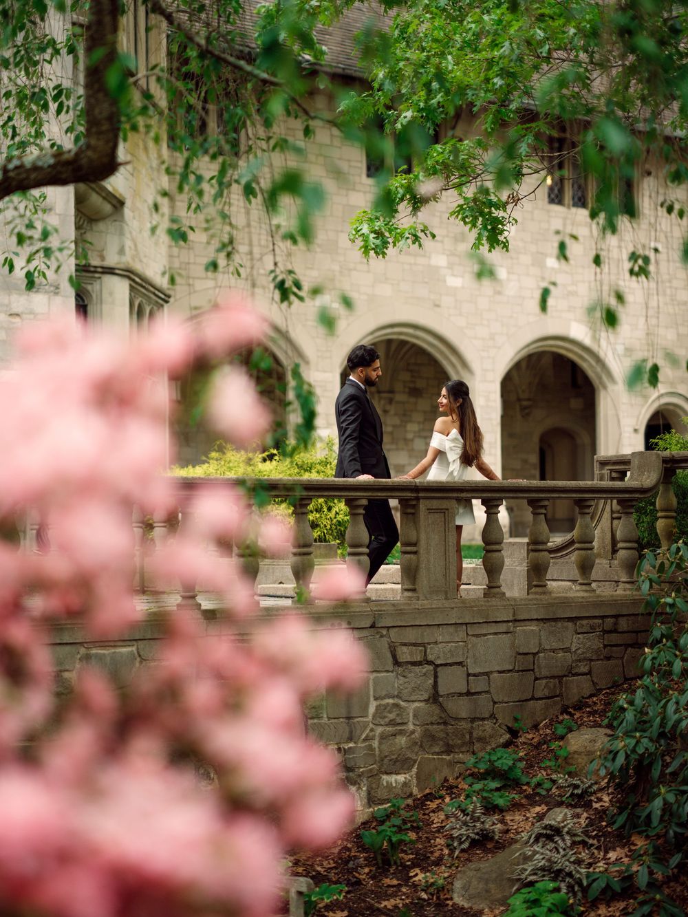 A romantic wedding photo at a stone archway with pink cherry blossoms in the foreground and green foliage overhead.