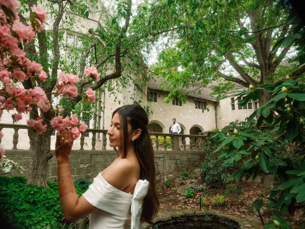 A woman in a white dress stands by blooming pink flowers in a garden with historic architecture visible behind.