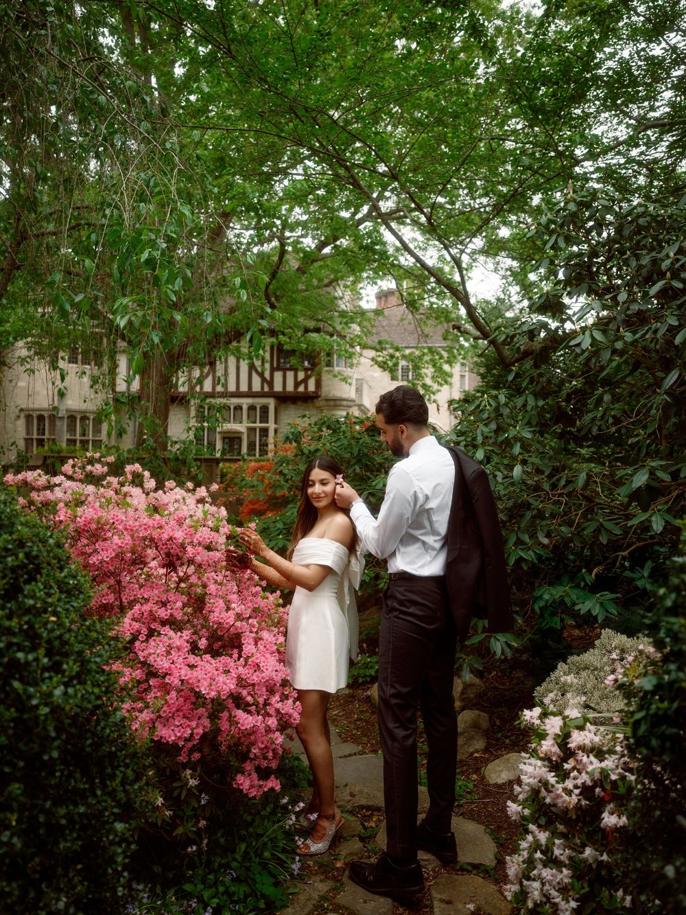 A romantic photo shoot in a garden with blooming pink azaleas and white flowers around a well-dressed couple.