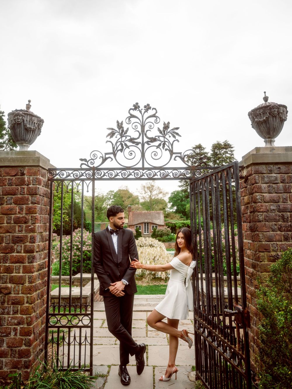 Elegant couple poses by ornate wrought iron garden gate with brick pillars and decorative finials at historic venue.