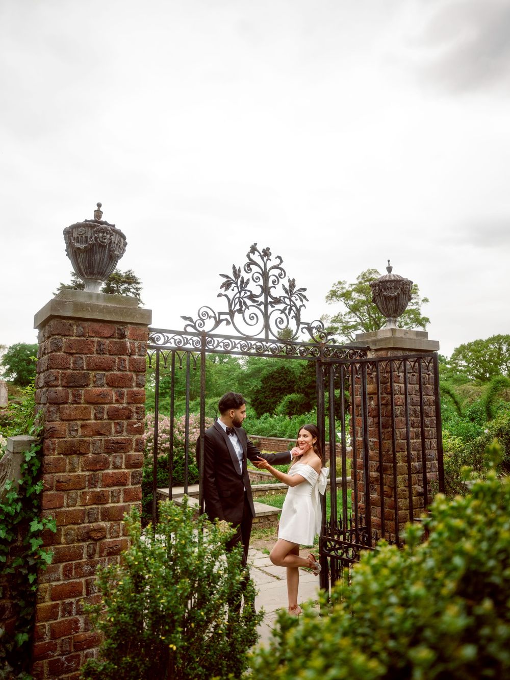 A pair shares a romantic moment by an ornate wrought iron gate with brick pillars in a garden setting.