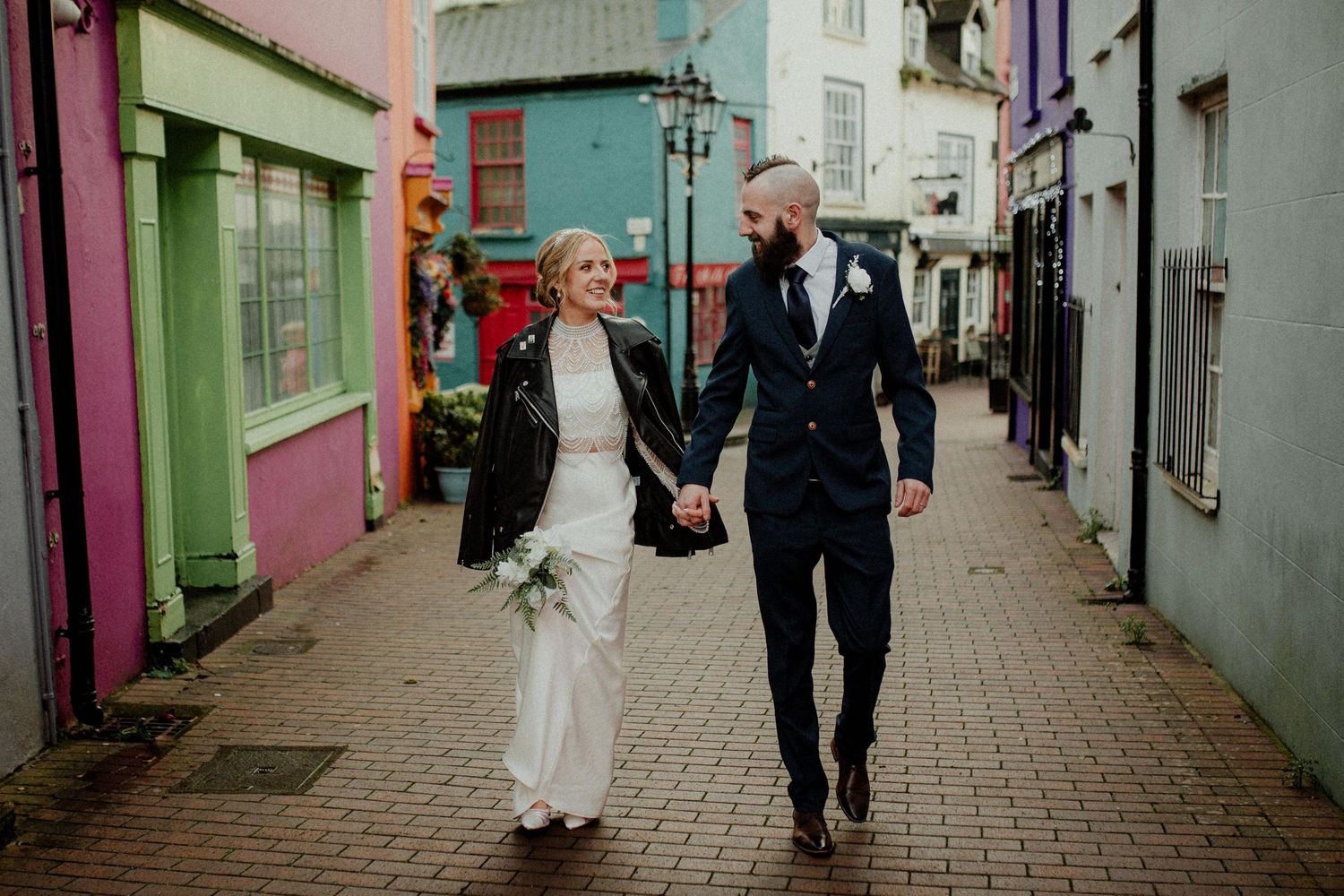 A couple walks down a charming colorful street with pink and blue buildings on their wedding day.