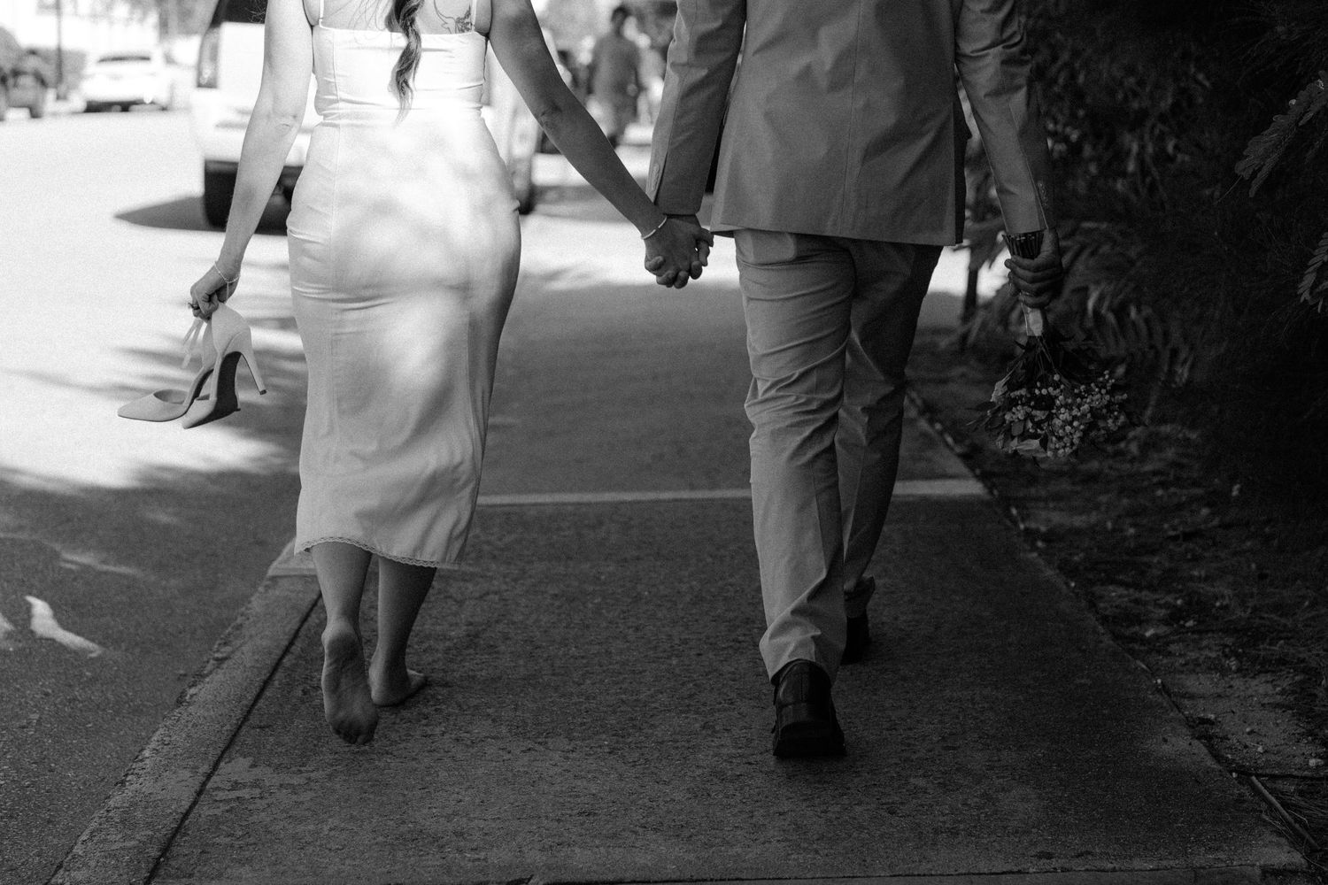 Black and white photo of a couple holding hands while walking together on a sidewalk path during a sunny day.