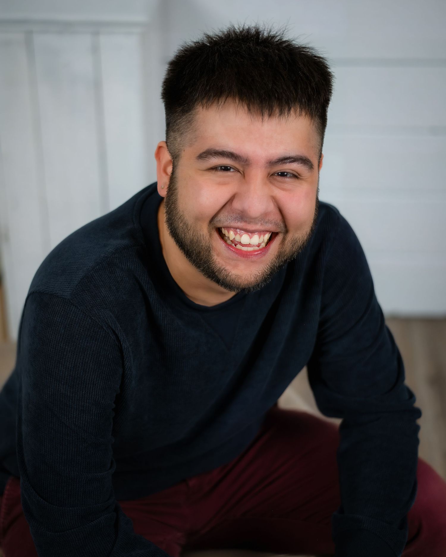 Smiling person wearing a navy sweater sits casually against white wall background.
