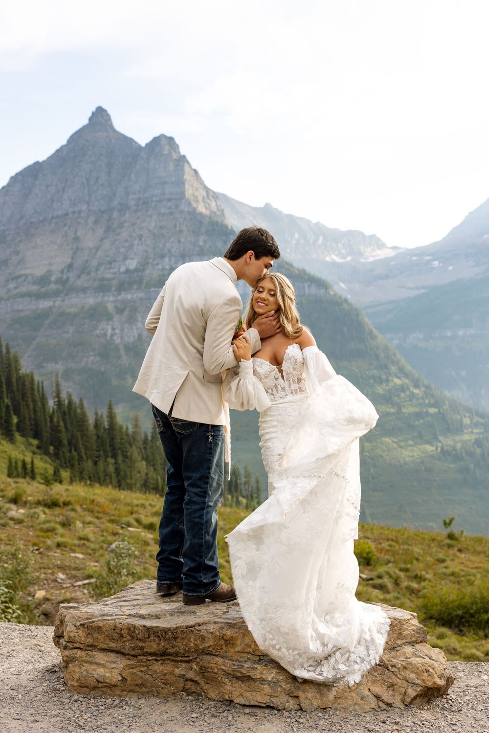 A romantic wedding couple stands on a rock with majestic mountain peaks of Glacier National Park in the background.