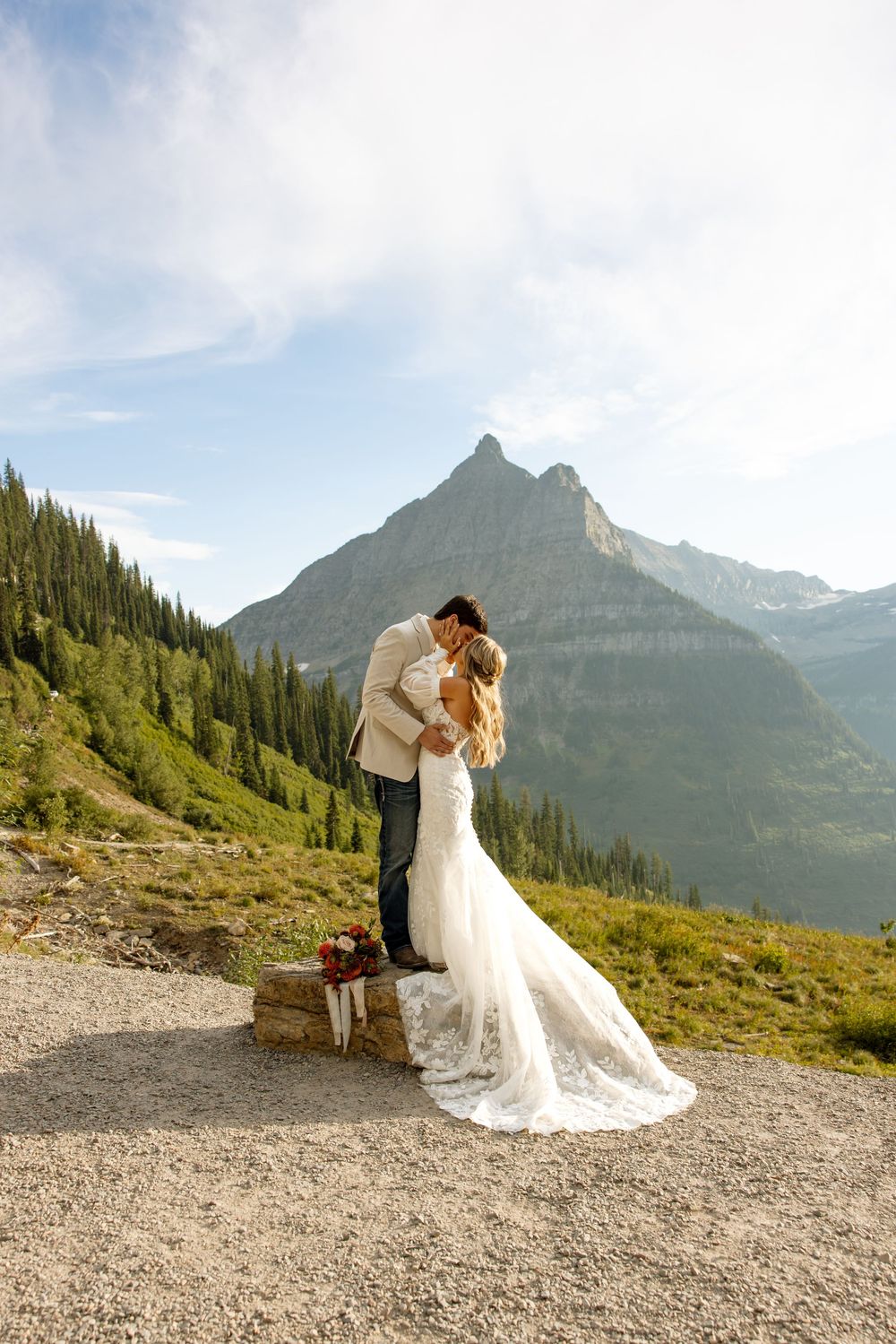 A couple in wedding attire embrace on a mountain trail with dramatic peaks and evergreen forests of Glacier National Park behind them.