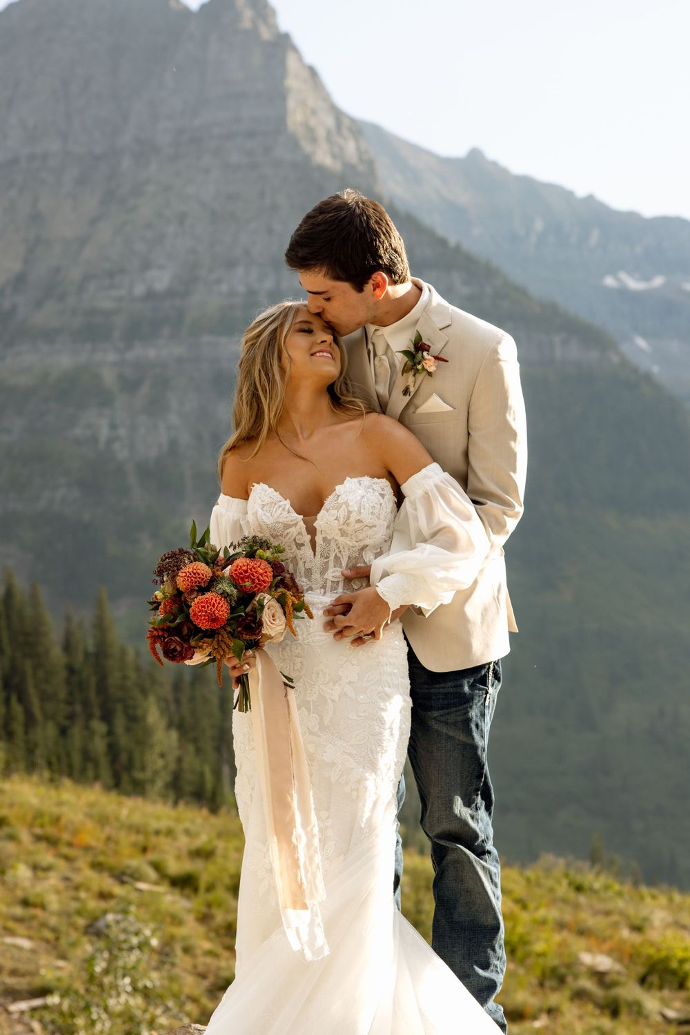 A romantic wedding couple embraces on a mountainside with dramatic rocky peaks and evergreen forest in the background.