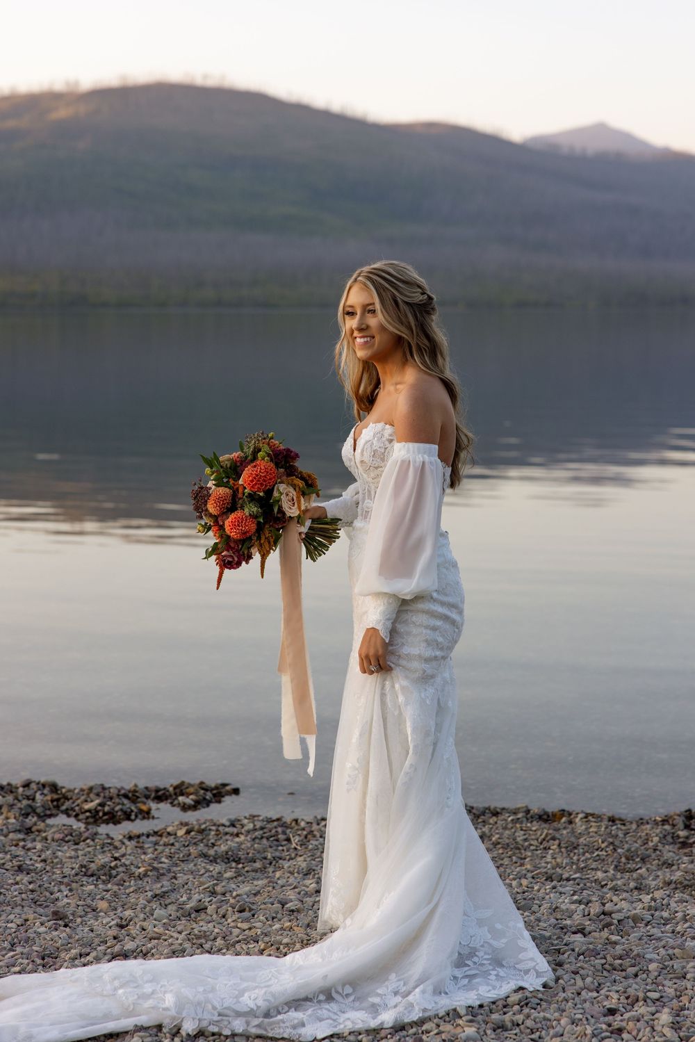 A bride in an off-shoulder wedding gown holds a red floral bouquet by a serene mountain lake at sunset.