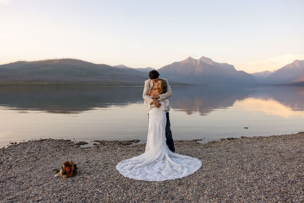 Two people embrace in elegant wedding attire on a rocky lakeshore with misty mountains and sunset reflections in the background.