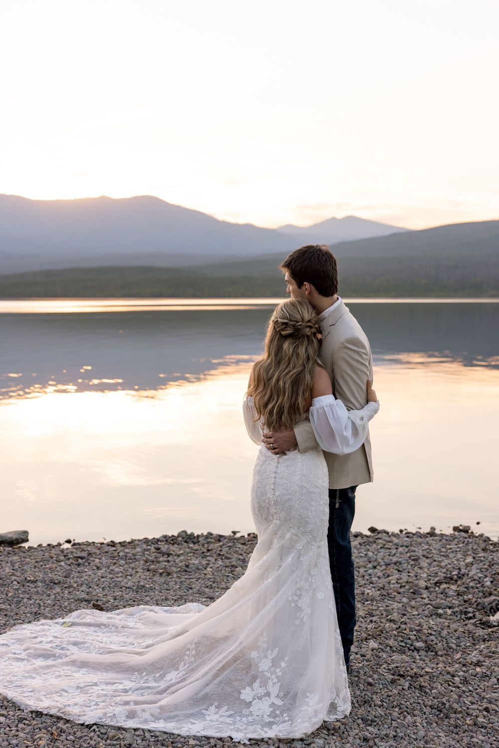 Couple embraces at sunrise by mountain lake shore with bride wearing elegant lace wedding gown with train.