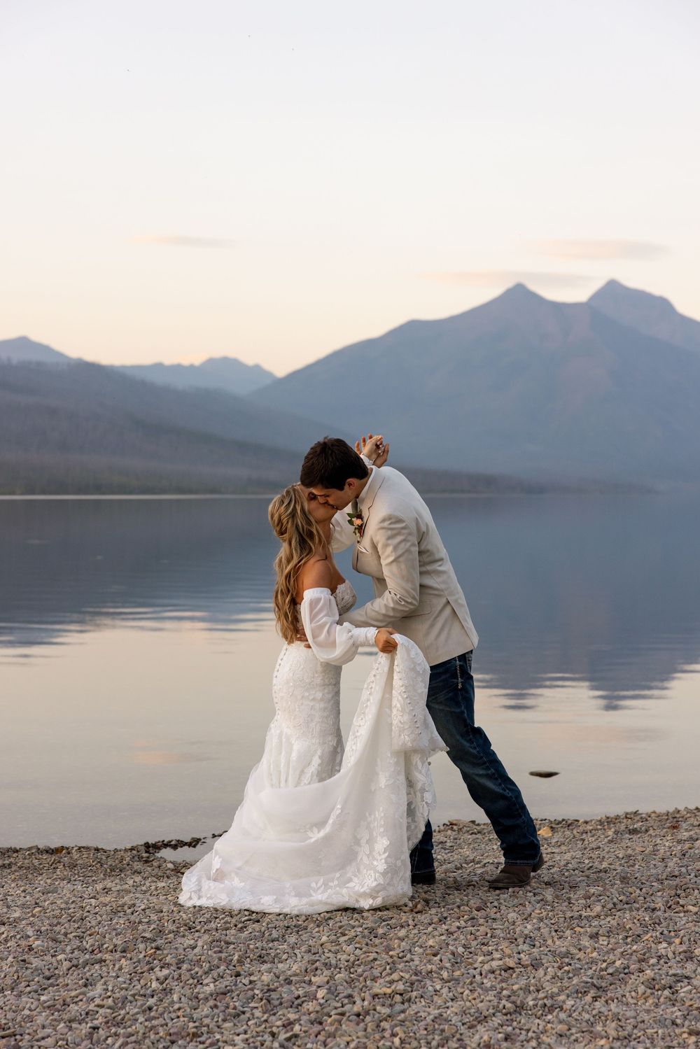 A romantic couple embraces on a rocky lakeshore with misty mountain peaks in the background during sunset.