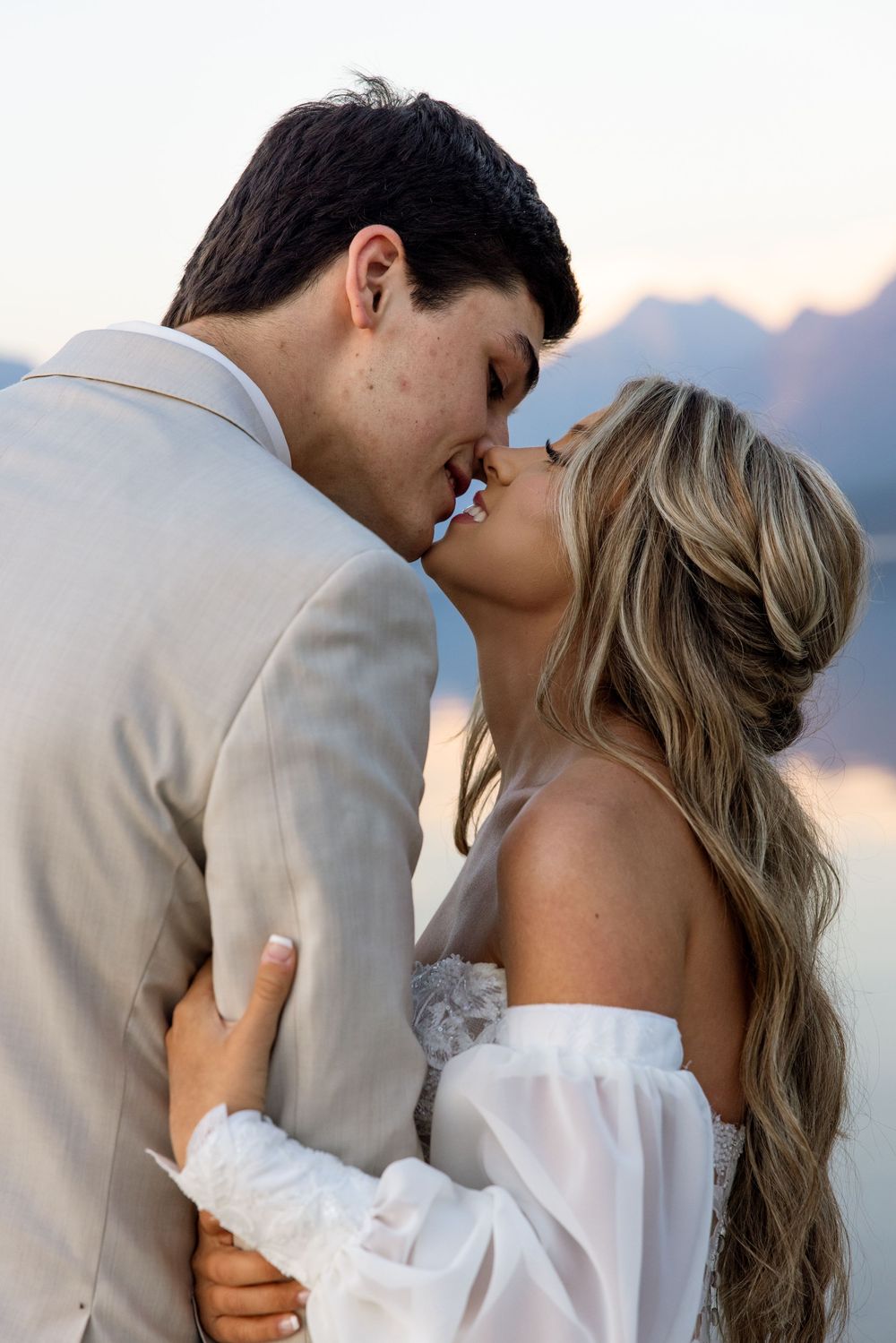 A romantic couple shares a tender kiss at sunset with mountains and water in the background.