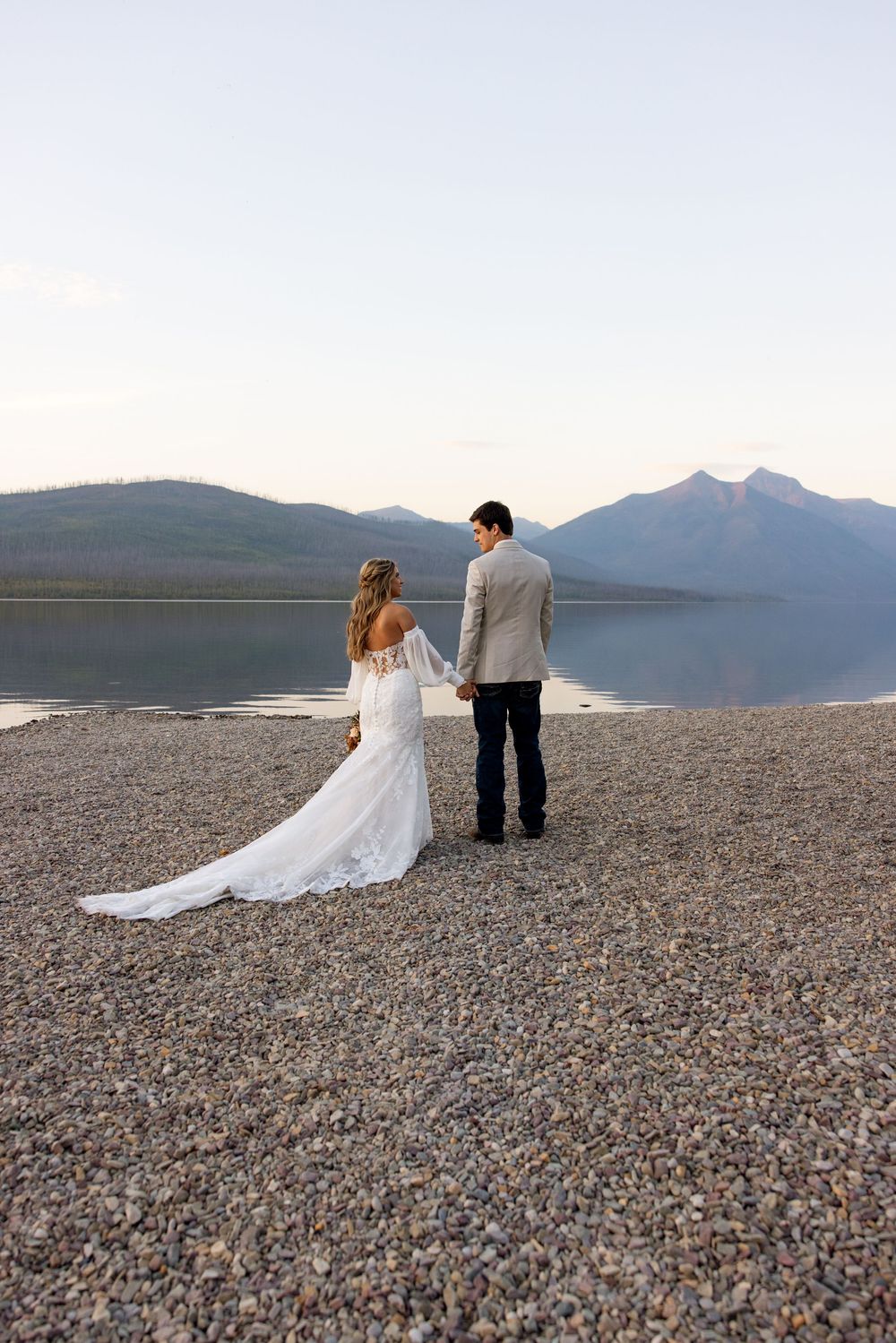 A couple in wedding attire stands on a rocky beach shore overlooking a serene mountain lake at dusk.