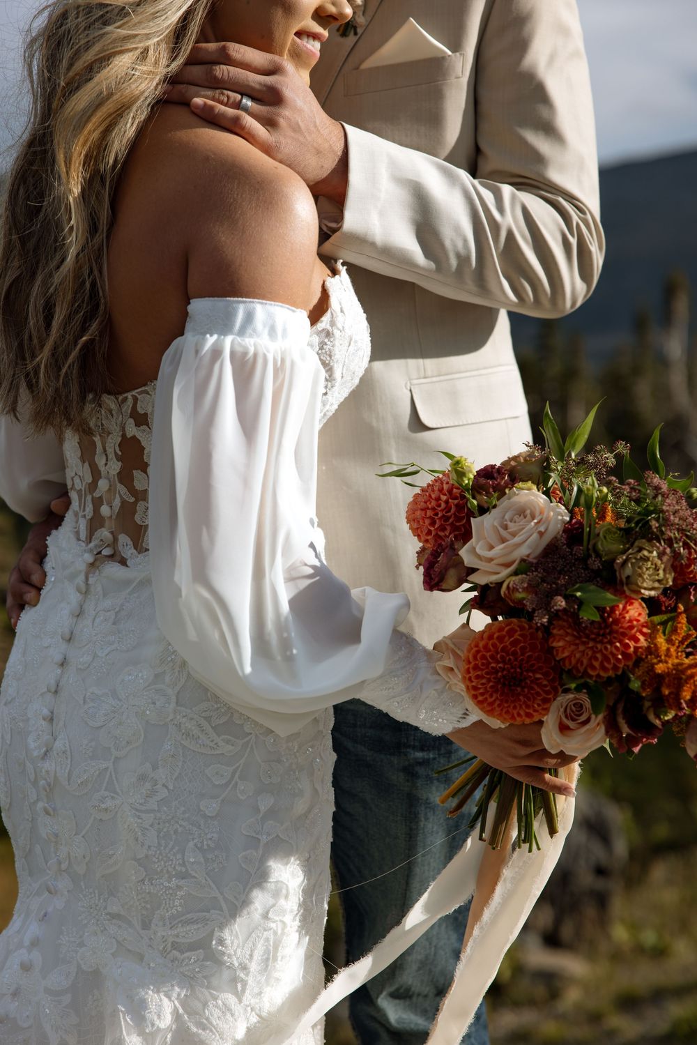 Close-up of bride in off-shoulder lace dress holding autumn-colored bouquet with red and pink florals.
