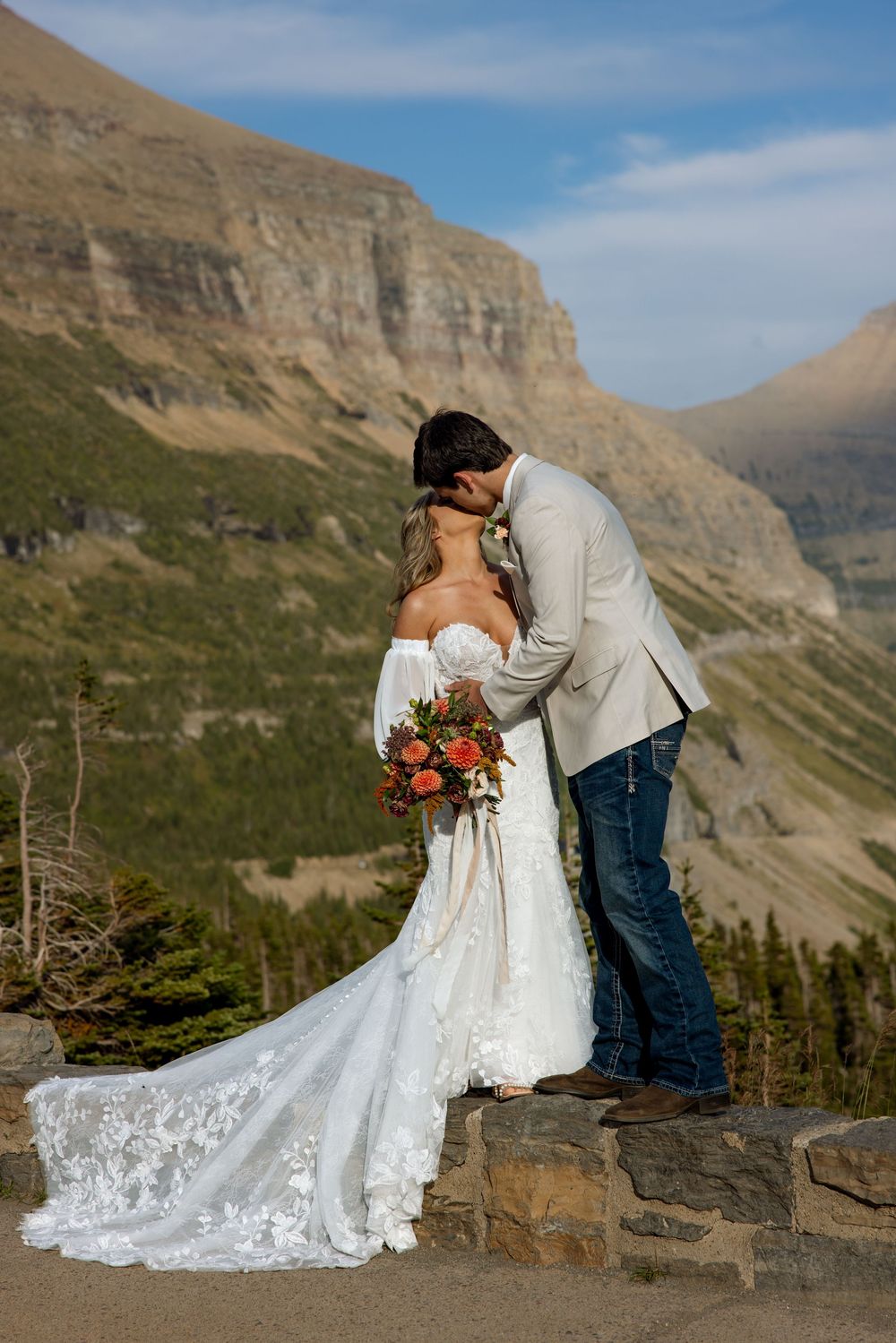 A couple shares an intimate wedding kiss on a stone wall overlooking dramatic mountain cliffs and valley in Glacier National Park.