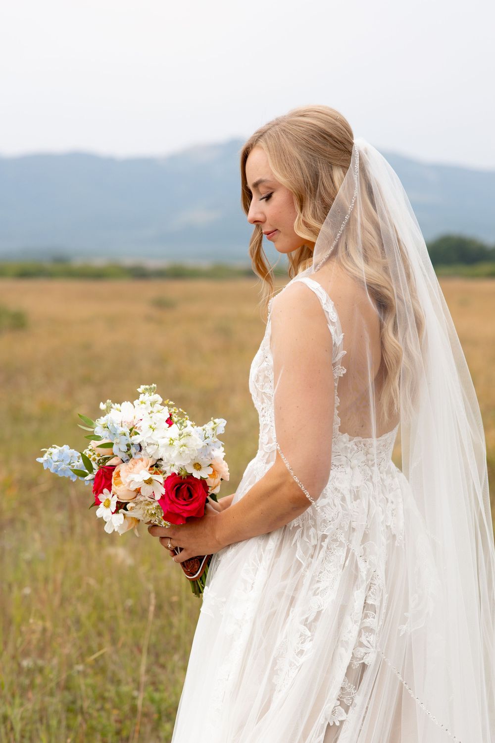 A bride in a lace wedding dress with long veil holds a vibrant bouquet of red and white flowers in a grassy meadow.