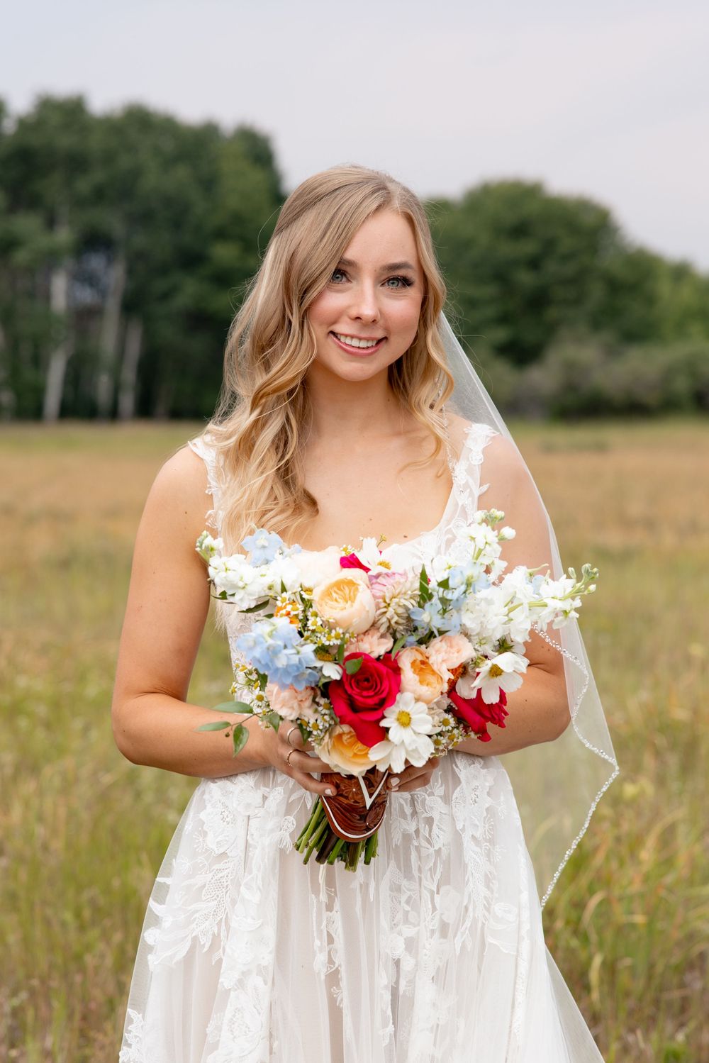 Beautiful bride wearing a white lace dress holds a vibrant bouquet of colorful flowers while standing in a meadow.