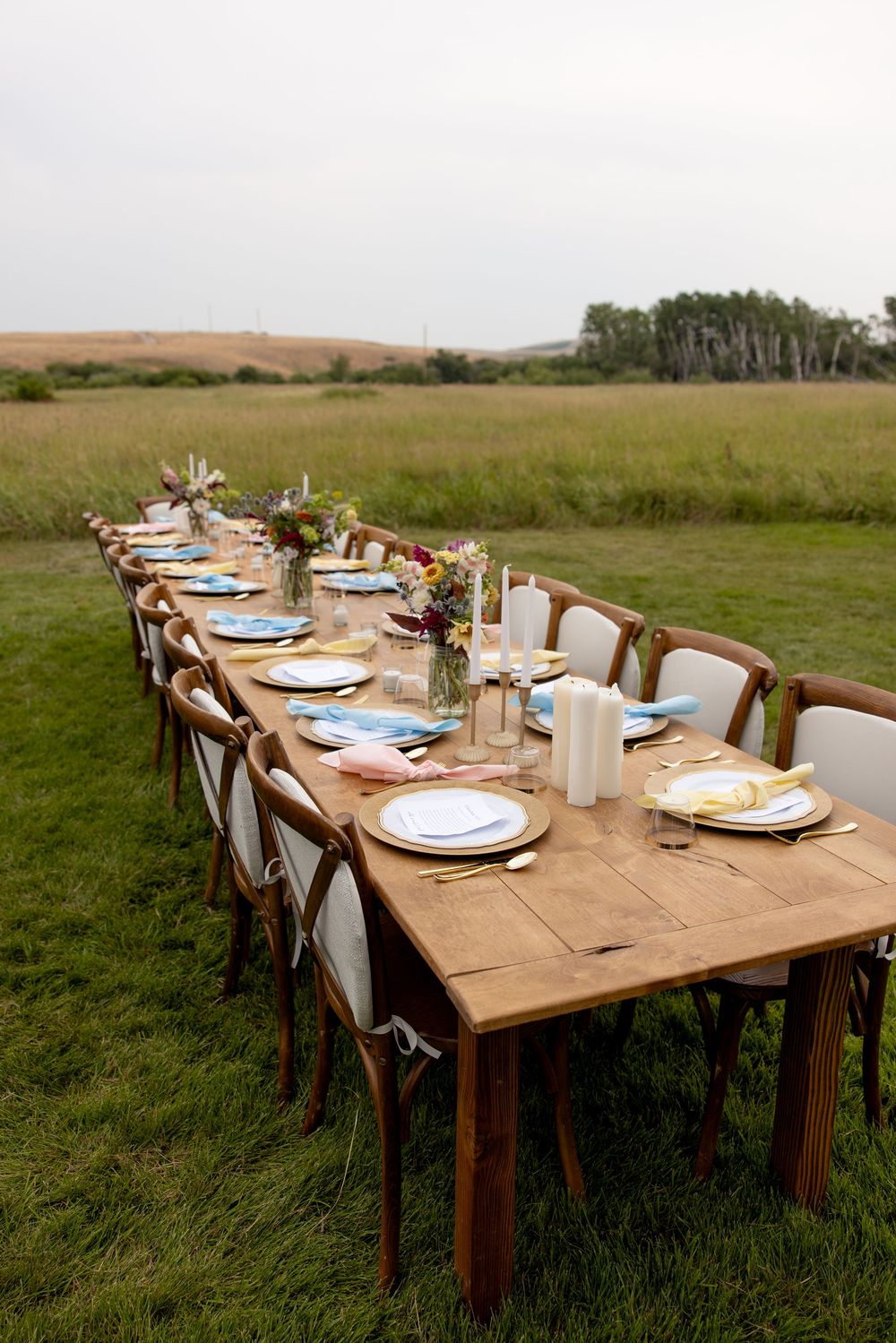 Long rustic wooden dining table set up outdoors with elegant place settings and floral centerpieces.