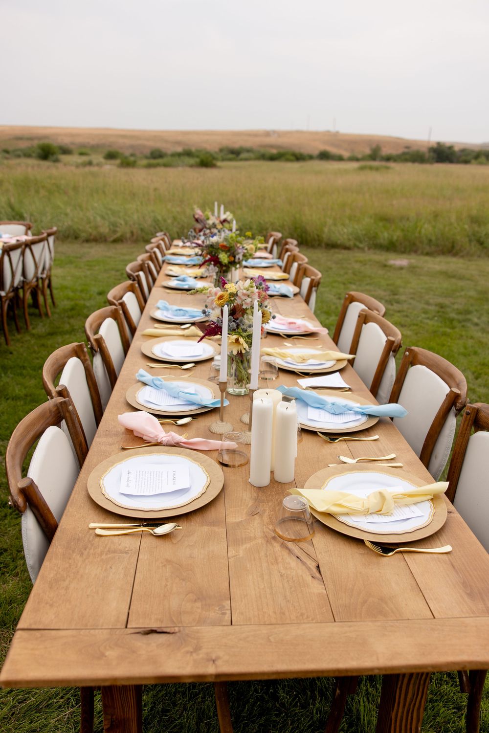 Rustic farm table set for outdoor dining with elegant place settings and floral centerpieces in a grassy field.