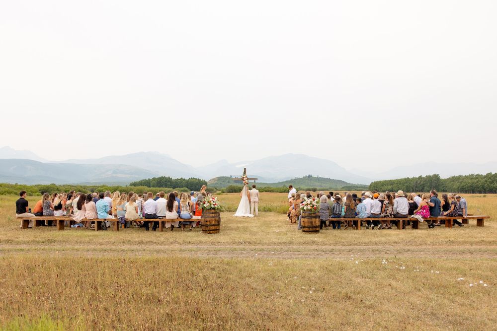 A scenic outdoor wedding ceremony in a golden field with mountain views as guests sit on wooden benches.