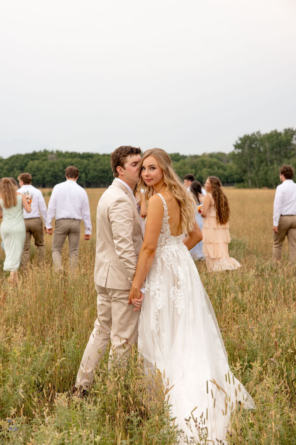 A couple in formal wedding attire poses together in a rustic field setting with their wedding party in the background.