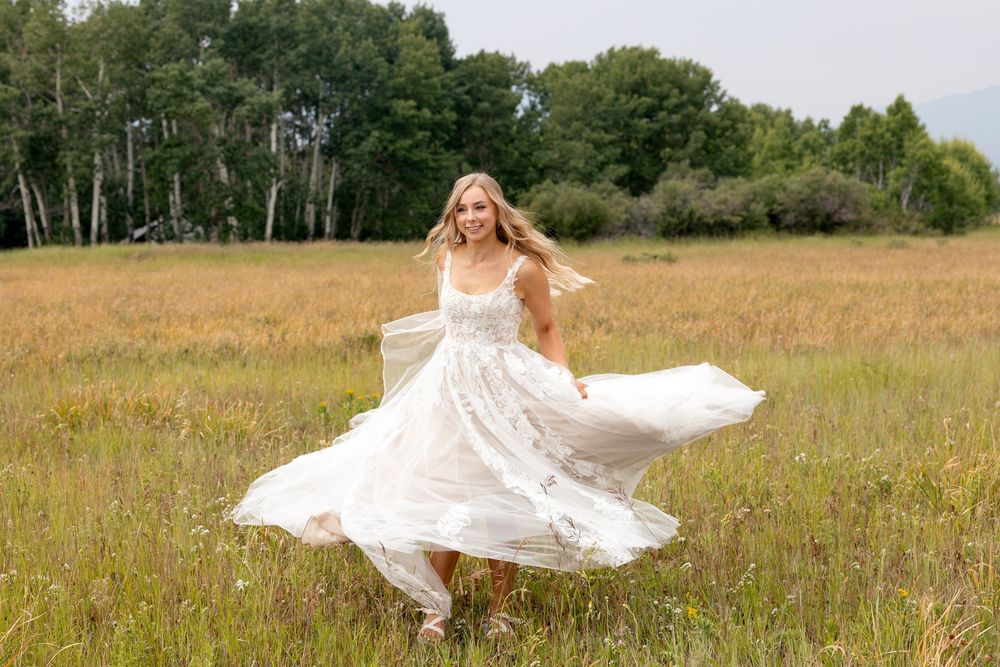 A white wedding dress twirls elegantly in motion against a sunlit summer meadow with trees in the background.