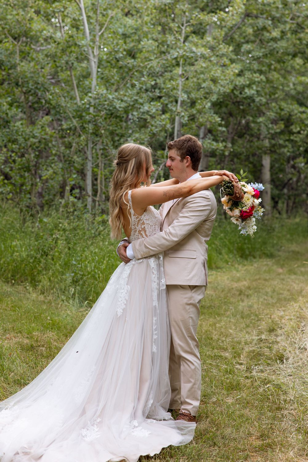A bride and groom share a romantic embrace in a woodland setting, with the bride holding a pink floral bouquet.