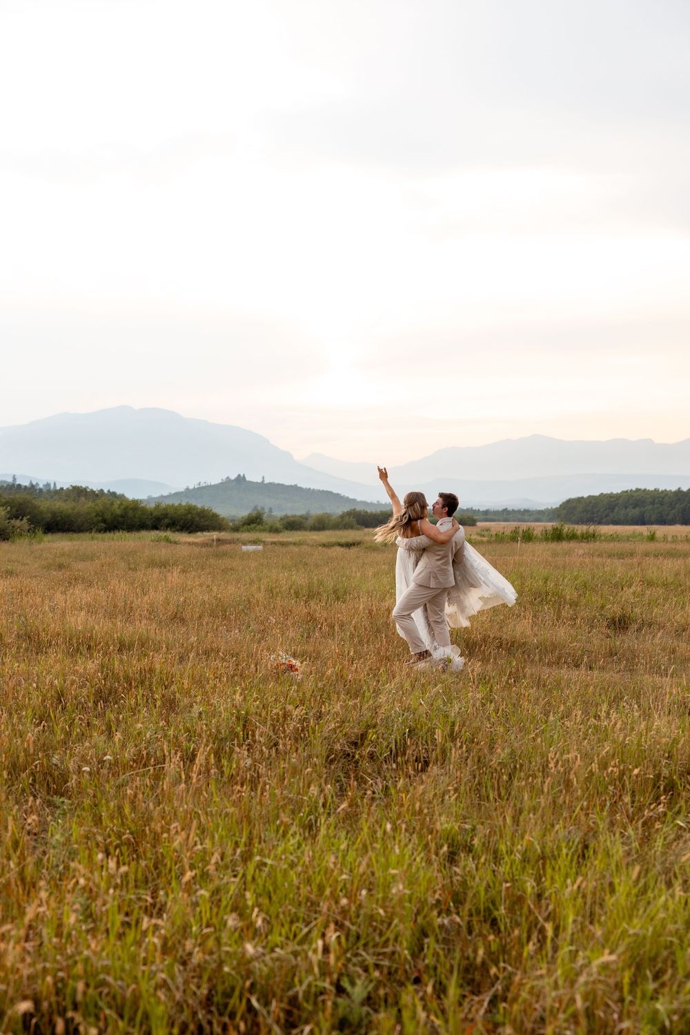 A couple dances joyfully in a sunlit meadow with mountains in the background during golden hour.