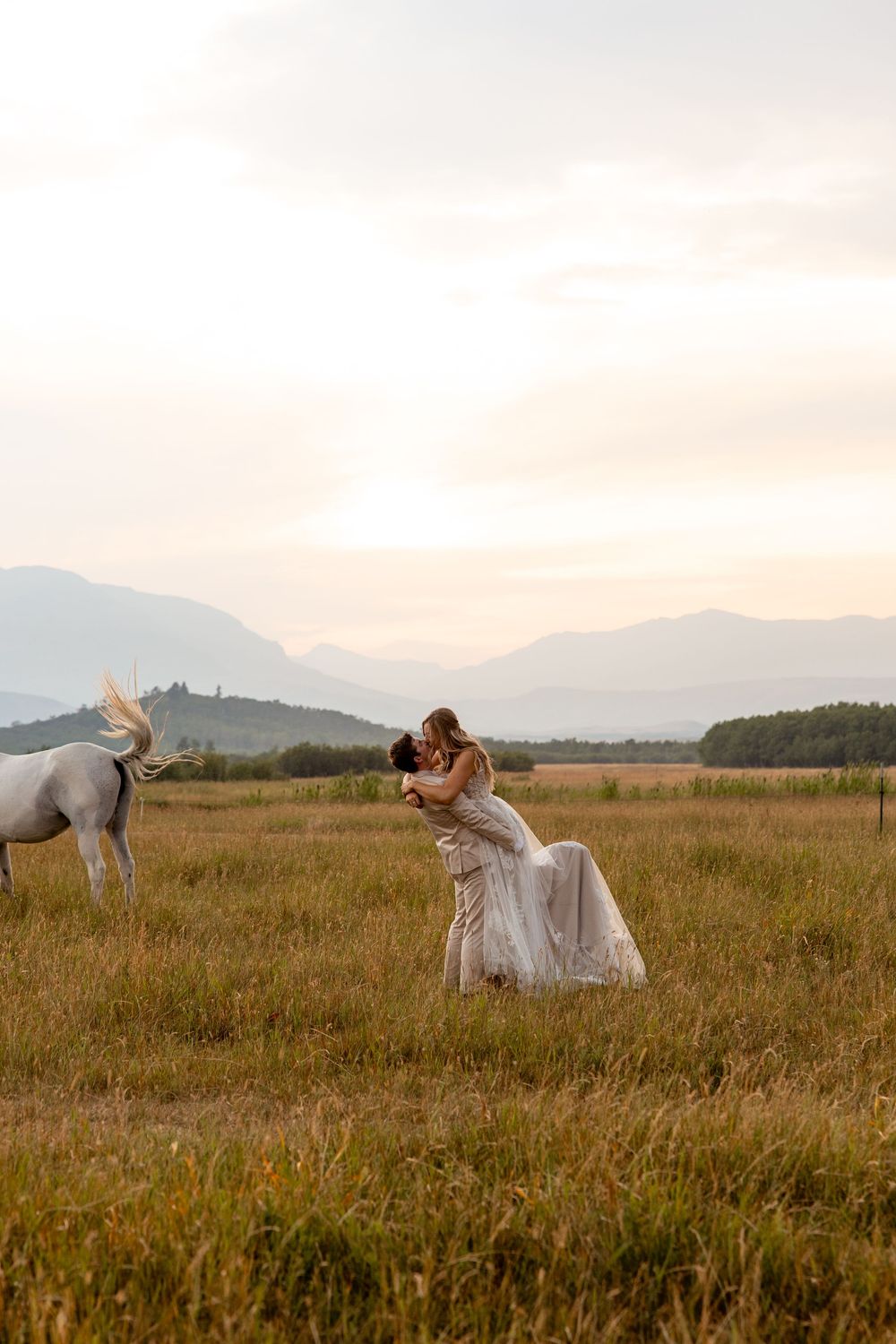 A white horse grazes in a mountain meadow at sunset while someone in a flowing white dress poses in the field.