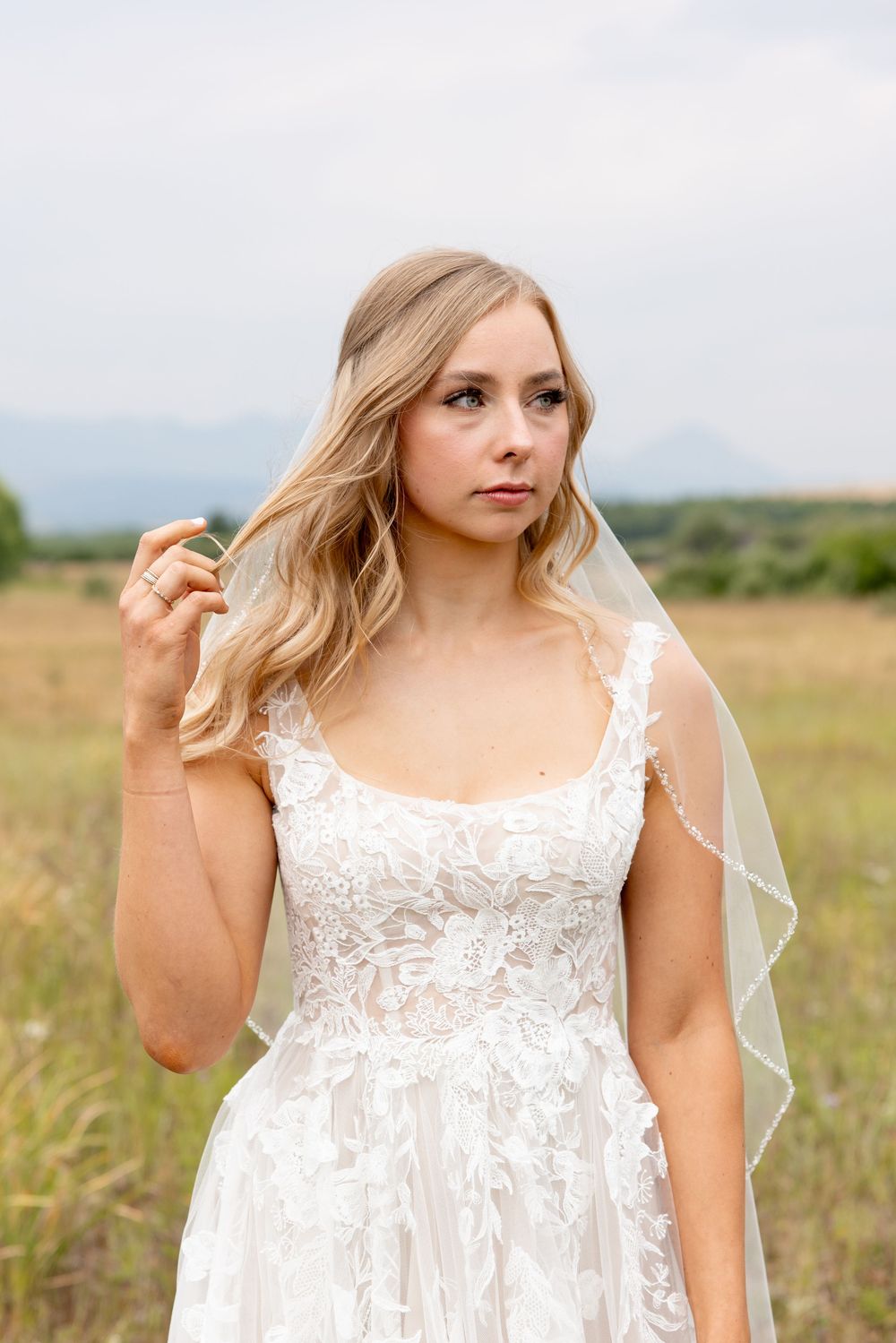 A bride in a white lace wedding dress with long wavy blonde hair stands in a sunlit meadow on an overcast day.