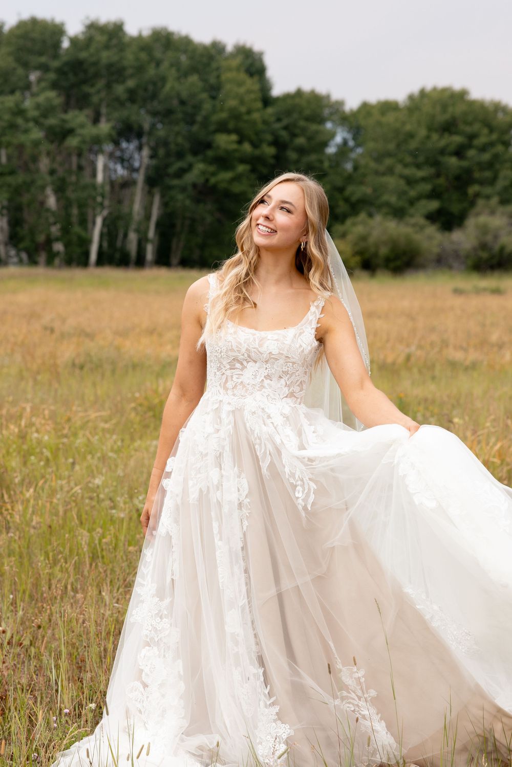 A bride in a flowing lace wedding dress twirls joyfully in a golden field with trees in the background.