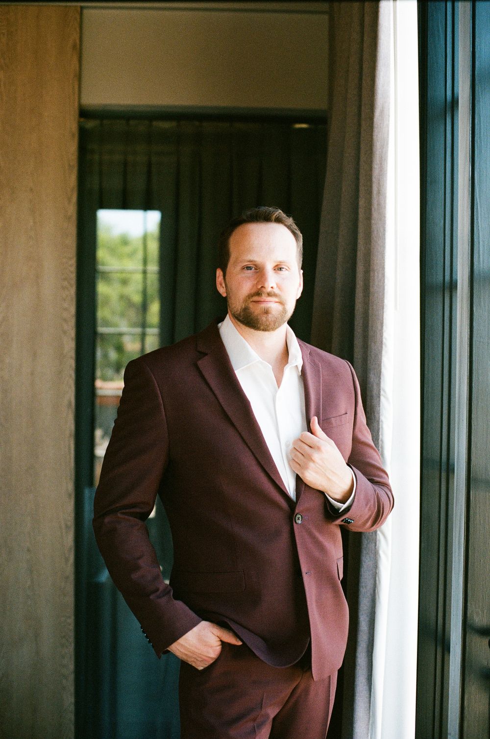 Handsome groom in maroon suit posing confidently by window with natural light on wedding day.