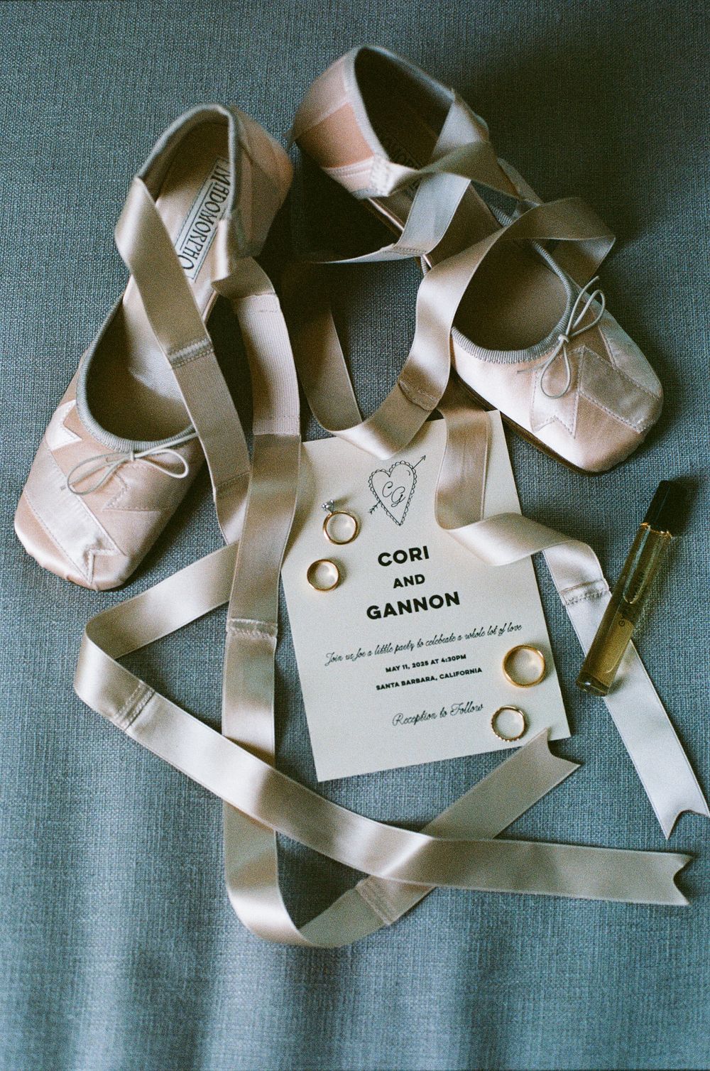 Wedding flat lay featuring ballet shoes, invitation for Cori and Gannon, gold rings, and a perfume bottle.