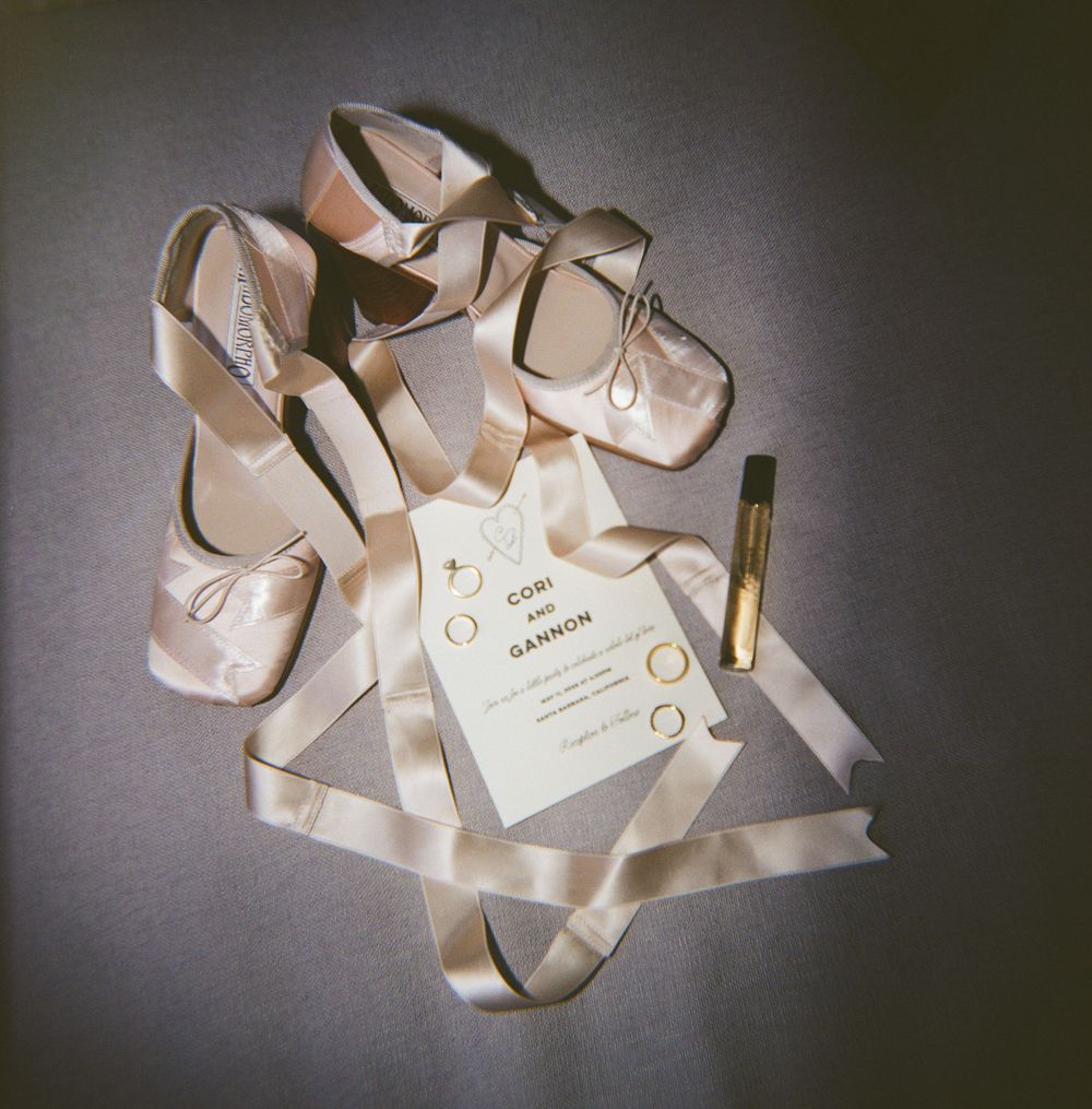 Elegant bridal flat shoes with satin ribbons beside a wedding invitation and gold perfume on gray background.