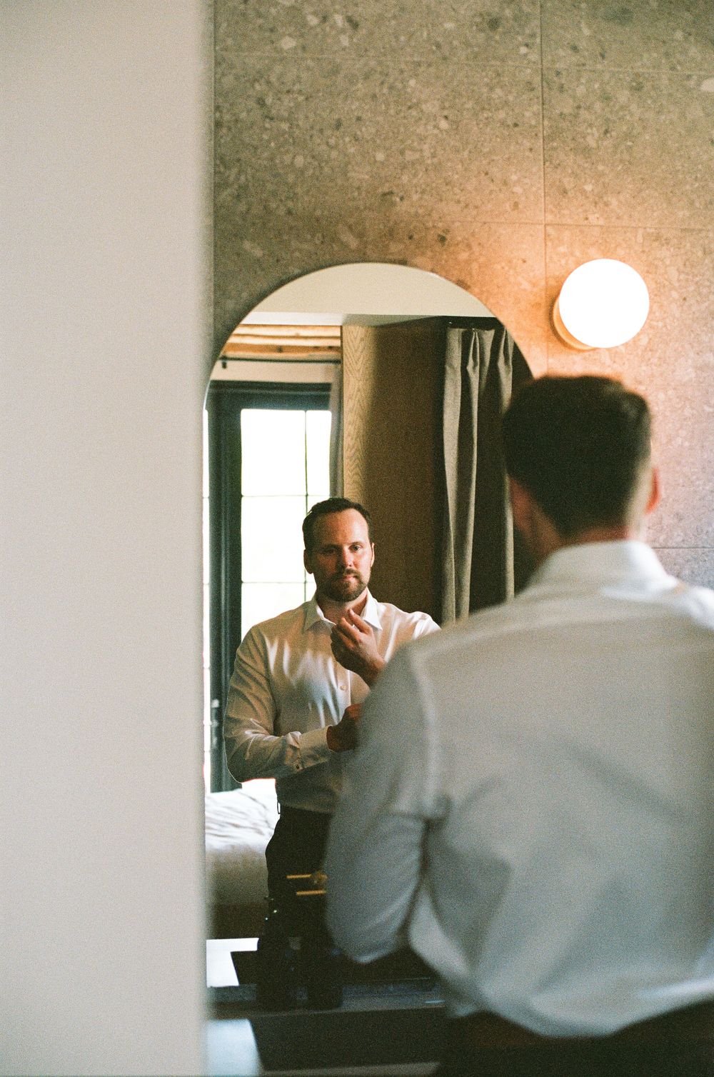 Groom in a white dress shirt adjusts his collar while looking at his reflection in an arched mirror.