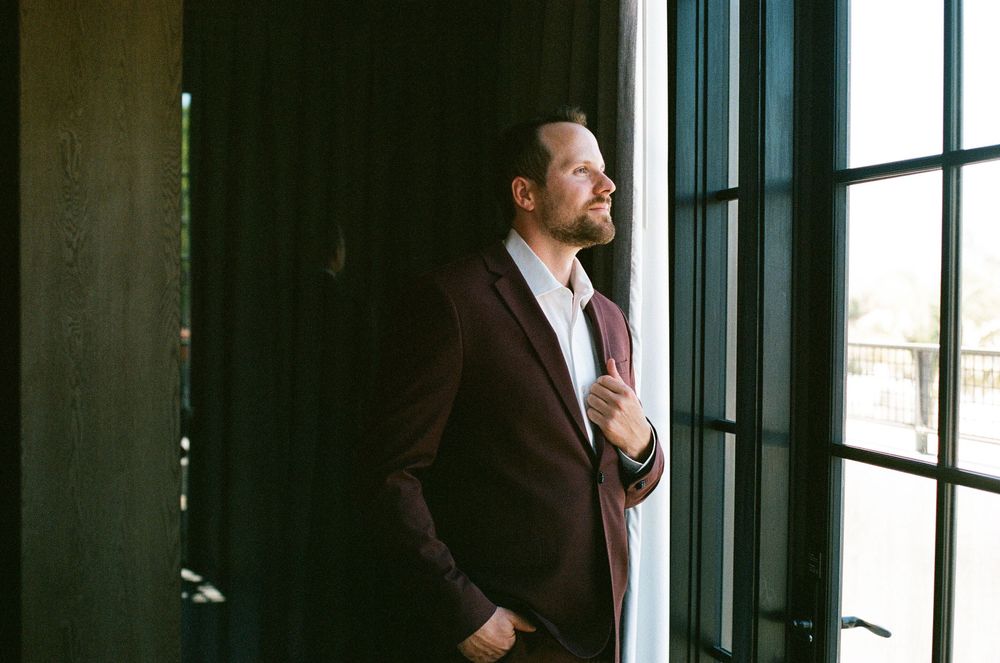 Groom in burgundy suit adjusting jacket while standing by dark French doors in soft light.