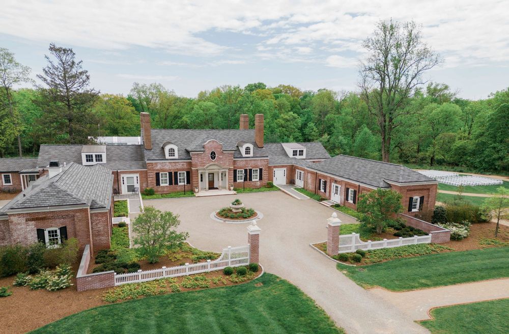 Aerial view of a luxurious brick mansion with circular driveway and manicured landscaping surrounded by lush trees.
