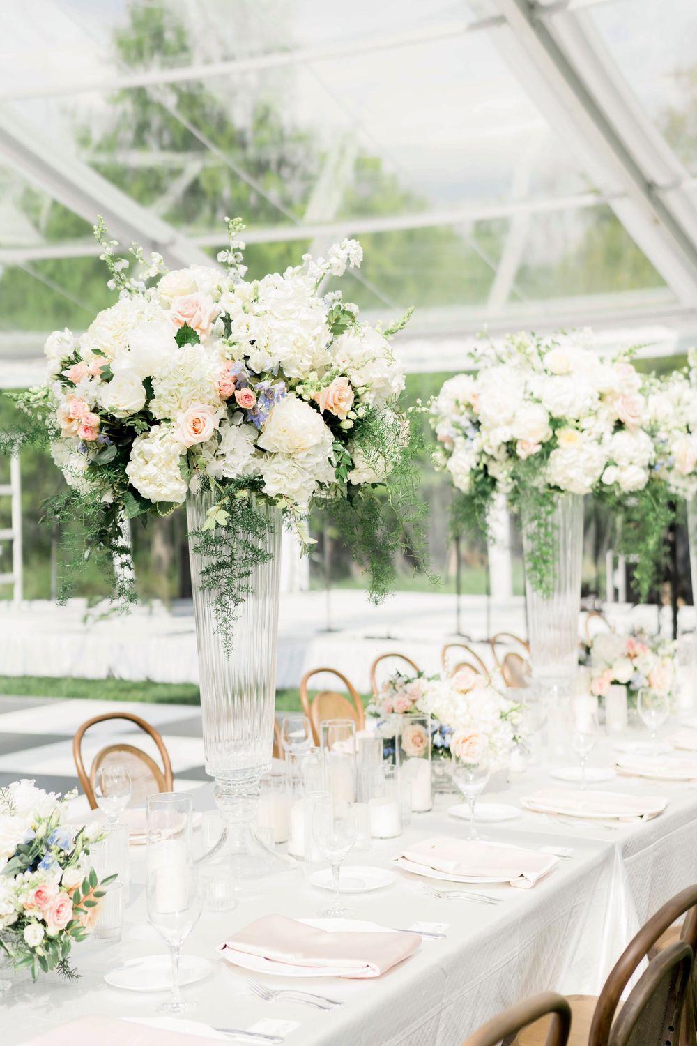Elegant white and blush floral centerpieces in crystal vases decorate a romantic wedding reception table under a clear tent.